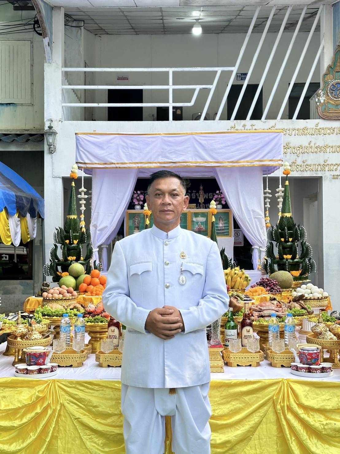 ชายสวมชุดขาว ยืนหน้าโต๊ะเครื่องเซ่นไหว้ในงานบุญ